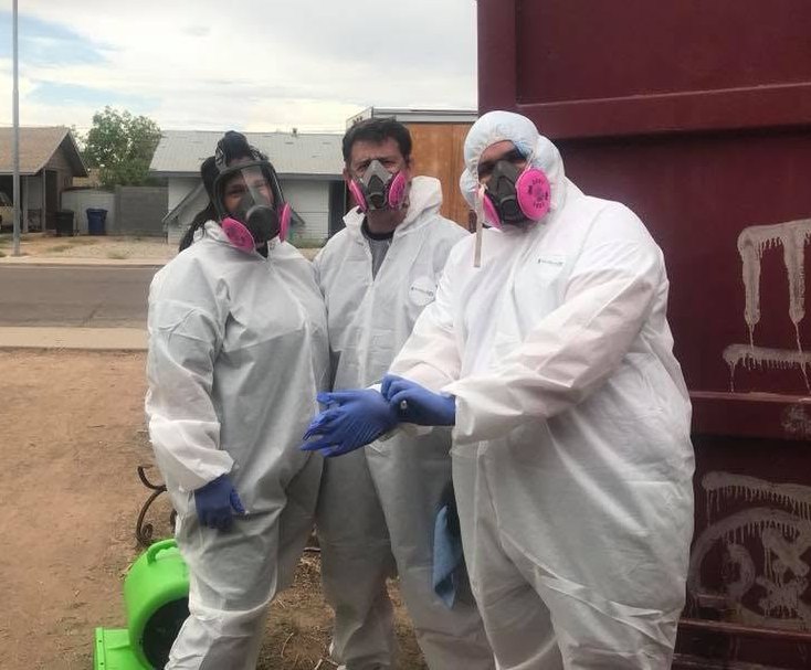 Three restoration workers in full Tyvek suits and respirators standing beside a dumpster and an air mover outside a residential property.